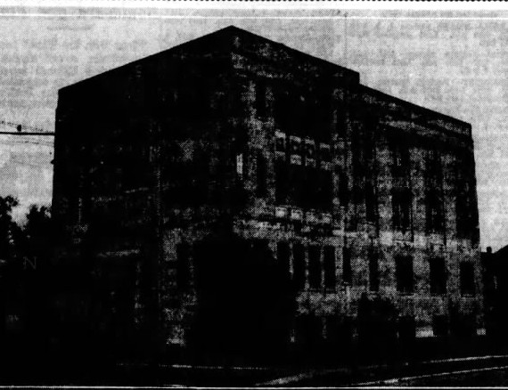 Dark newspaper photograph, 1943: a boxy three-story building seen at an angle from the corner, with rows of windows, trees at left and an overhead wire.
